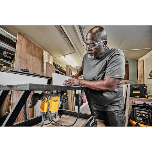 A person working in a workshop is using a DEWALT router mounted on a workbench to shape a piece of wood. The individual’s face is blurred for privacy. Various DEWALT toolboxes and woodworking materials are visible in the background.