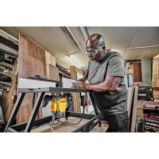 A person working in a woodworking shop is using a DEWALT DWE627KT router mounted on a table to shape a piece of wood. Various wooden panels and tools are visible in the background.
