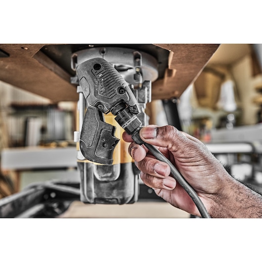 Close-up of a hand connecting a power cord to a DEWALT DWE627KT woodworking tool mounted under a workbench, in a workshop setting.