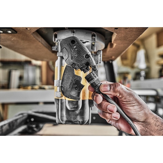 Close-up of a hand connecting a power cable to a DEWALT router tool mounted under a workbench in a workshop setting.