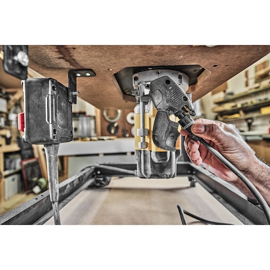 A close-up view of a hand connecting a power cable to a DEWALT router mounted underneath a workbench in a workshop.
