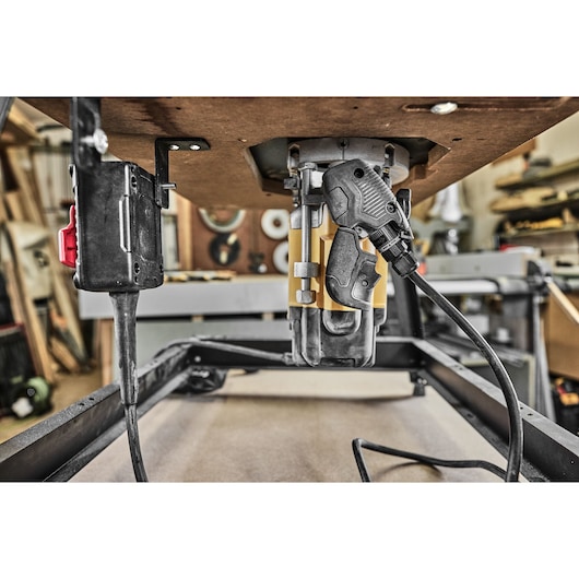 Close-up image showing the underside of a workbench with a DEWALT router mounted upside down and connected to a power source. The photo is taken inside a woodworking workshop, with tools and materials visible in the background.