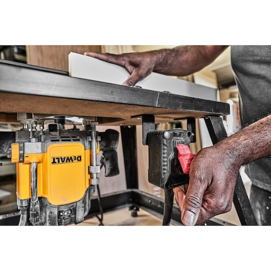 A person operates a DEWALT router mounted underneath a workbench, adjusting the switch with one hand while guiding a piece of material on top of the bench.