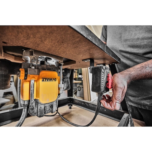 A close-up view of a yellow DEWALT router mounted underneath a work table. A person is turning on the power switch attached to the table, preparing the tool for use.