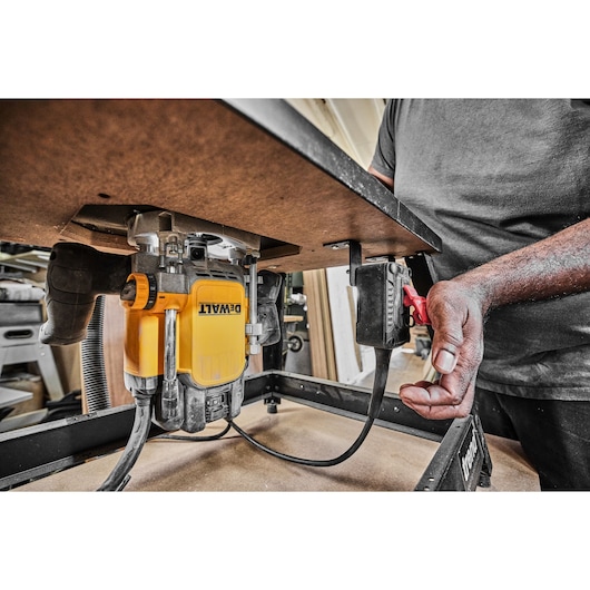 A close-up view of a DEWALT router mounted underneath a work table, with a person operating a power switch attached to the table. The workspace includes workshop equipment and materials in the background.