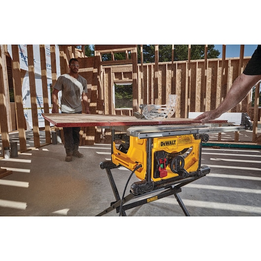 A yellow DEWALT table saw is cutting a piece of wood inside a partially constructed wooden building. One person is operating the saw, and another stands in the background. Sawdust is visible around the machine.
