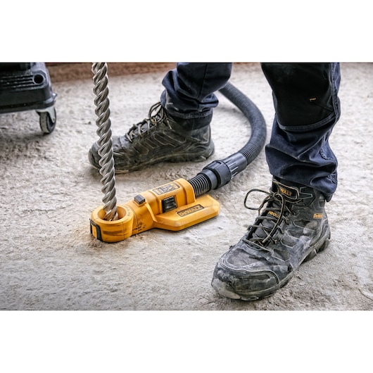 A close-up of a DEWALT DWH050 dust extraction attachment connected to a large drill bit and hose, being used on a concrete surface by someone wearing safety boots and work pants.
