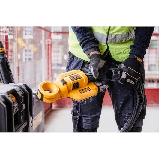 A person wearing safety gloves and a reflective vest uses a DEWALT DWH050 tool attachment for dust collection at a construction site.