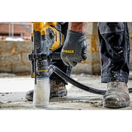 A close-up of a construction worker using a DEWALT power tool with a dust extraction attachment on a concrete floor. The worker is wearing gloves and work boots, and the environment appears to be a building site.