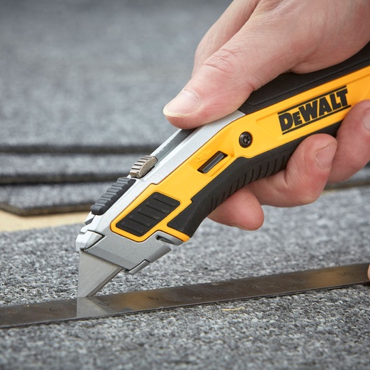 A close-up of a hand using a DEWALT utility knife to cut carpet along a metal ruler.