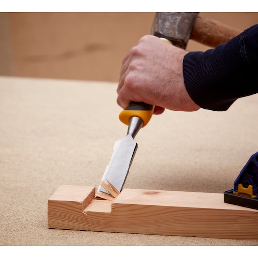 Hand using a DEWALT chisel and hammer to carve wood on a workbench.