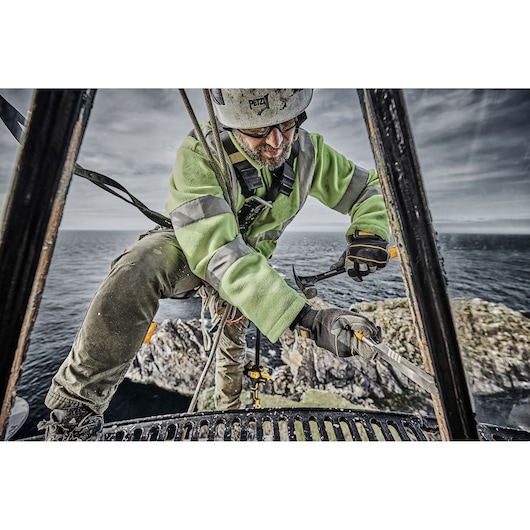 Worker in safety gear using a DEWALT tool outdoors near the sea, standing on metal scaffolding.