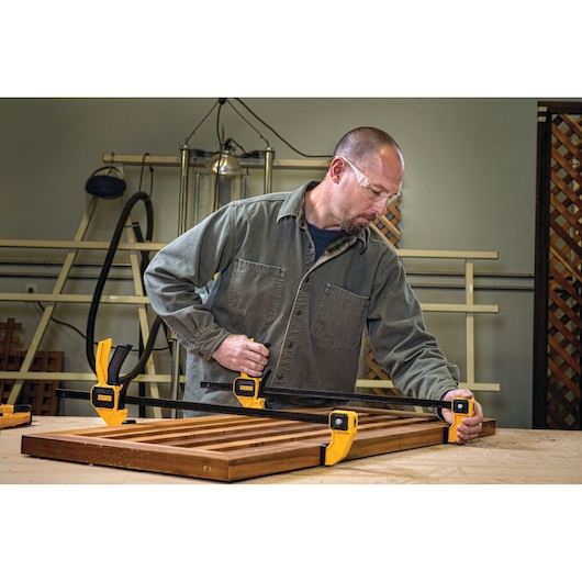 Person using DEWALT bar clamps to secure a wooden panel on a workbench in a workshop.
