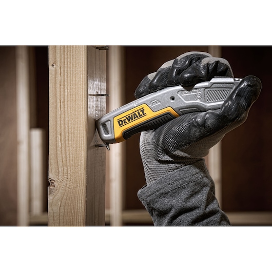 Gloved hand using a DEWALT utility knife against a wooden beam at a construction site.