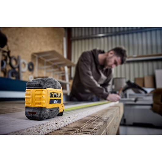 DEWALT 8M tape measure on a wood surface, person measuring in the background inside a workshop.