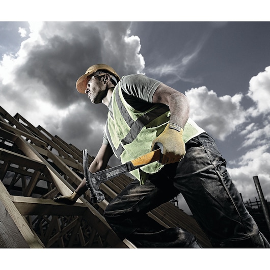 A construction worker wearing a safety vest and helmet is working on a wooden structure outdoors. The worker is holding a DEWALT hammer, and is wearing protective gloves. The sky is cloudy in the background.