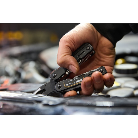 Hand uses DEWALT black multi-tool pliers, with various tools visible, in a workshop setting.