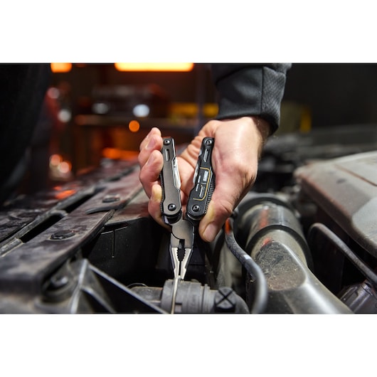 Hand using a DEWALT multitool pliers to grip a wire inside a car engine compartment.