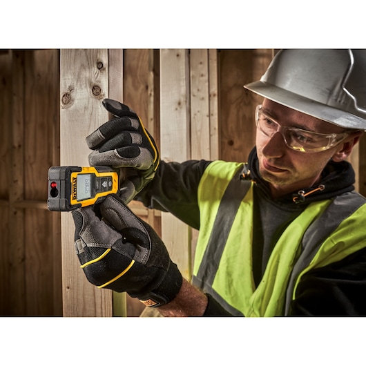 A person wearing gloves and a safety vest is using a DEWALT laser distance measurer (model DWHT77100) to measure wooden beams at a construction site. The person's face is blurred for privacy.