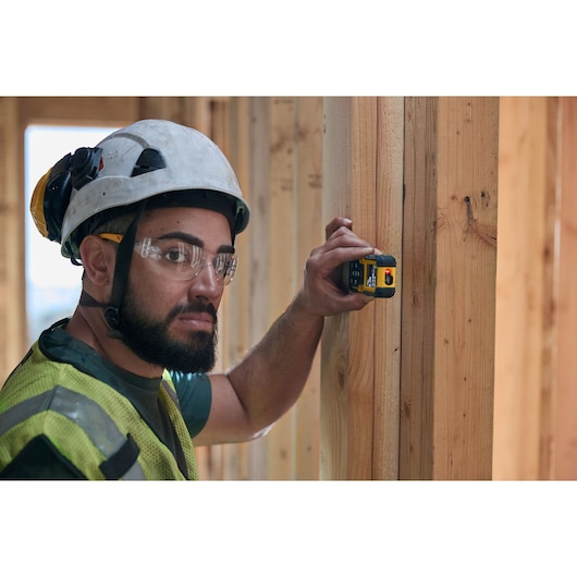 A construction worker wearing a safety helmet and reflective vest is using a DEWALT DWHT77200 laser distance measurer against a wooden beam inside a building under construction. The worker's face is obscured.