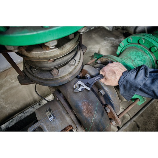A person using a DEWALT adjustable wrench (DWHT80268-0) to tighten a pipe fitting in an industrial setting with metal pipes and bolts.