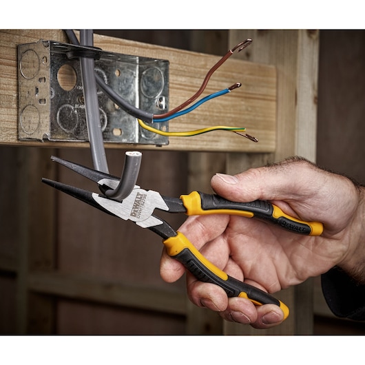 Hand using DEWALT pliers to cut electrical cable near an outlet box with exposed wires, shown during installation.