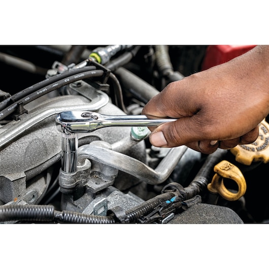 A close-up of a hand using a chrome ratchet wrench on a bolt inside a car engine compartment.