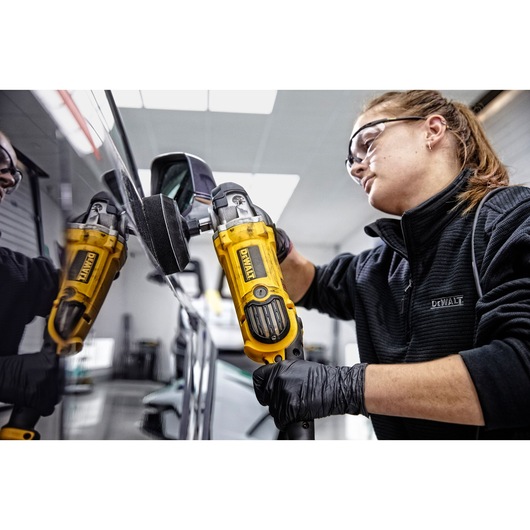 A person wearing black gloves and a black jacket is using a yellow DEWALT DWP849X polisher on the surface of a vehicle, with their face blurred.