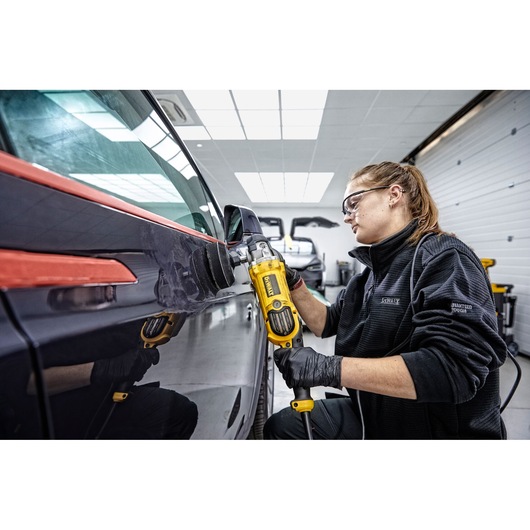 Person using a DEWALT DWP849X polisher on a car surface in a workshop. The individual is wearing black gloves and a dark jacket, and the setting is a brightly lit garage.