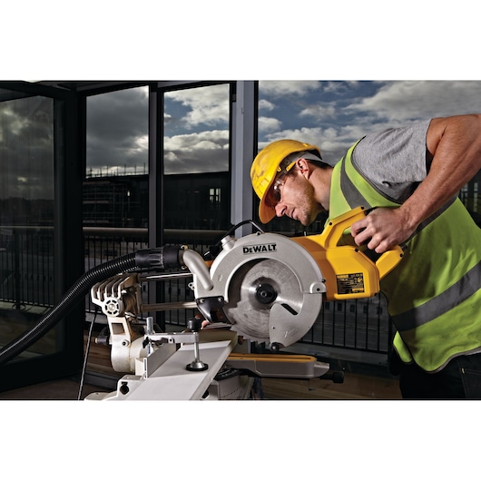 A person wearing a yellow hard hat and reflective vest operates a DEWALT DWS778 miter saw indoors, cutting a piece of material on a workbench. The background shows large windows with an outdoor view.