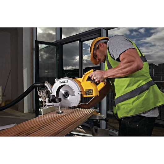 A person wearing a safety helmet and high-visibility vest is using a DEWALT DWS778 circular saw to cut a wooden plank indoors. The saw is connected to a dust extraction system. Large windows and construction materials are visible in the background.