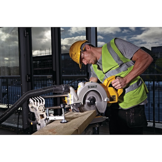 A person wearing a high-visibility vest and a hard hat is using a DEWALT miter saw to cut a wooden plank at a construction site. The scene is indoors with large windows showing an outdoor view.