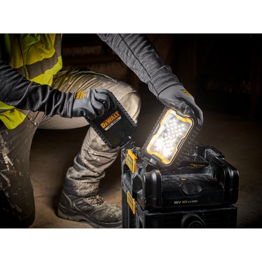 A person wearing work gloves and a yellow safety vest is adjusting a DEWALT portable work light, which is illuminated and placed on top of a black DEWALT tool box. The scene suggests a construction or workshop environment.