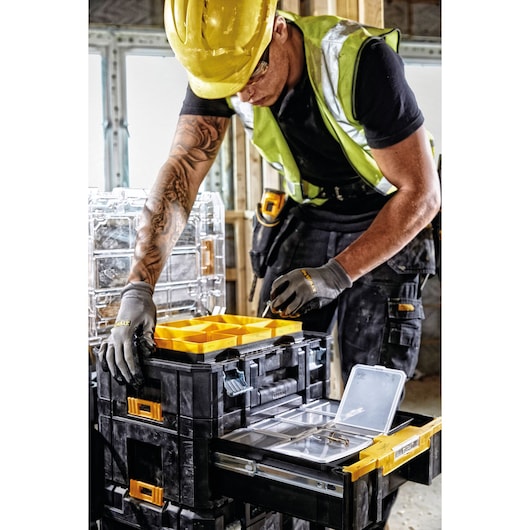 A construction worker wearing a yellow helmet and safety vest organizes tools and small parts in a DEWALT DWST1-70705 modular storage box system. The worker uses gloves and is arranging items in the yellow compartment trays and clear-lidded drawers of the black and yellow storage units, situated in a construction site.