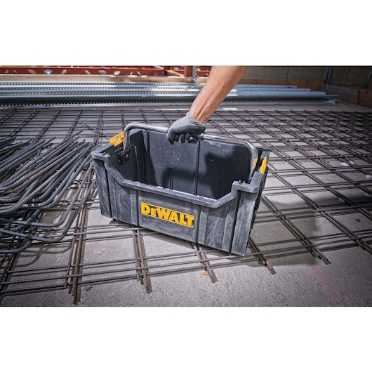 A DEWALT branded black plastic toolbox being carried by a person wearing a work glove, placed on a construction site with metal rebar grids.