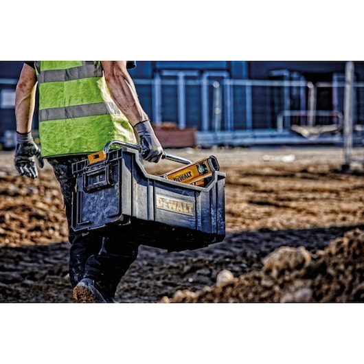 A person wearing a safety vest and gloves is carrying a black DEWALT toolbox with a handle and DEWALT tools inside, walking across a construction site.