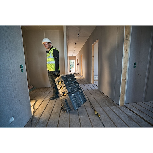 A worker wearing a high-visibility vest and hard hat is pulling a DEWALT DWST1-75799 wheeled tool storage box through an unfinished building corridor with wooden floors and drywall.