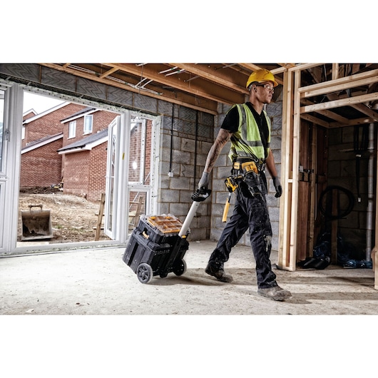 A person wearing a yellow hard hat, safety vest, gloves, and work trousers is pulling a DEWALT DWST1-75799 wheeled toolbox inside a construction site with unfinished walls and visible timber framing. The toolbox has black and yellow compartments and sturdy wheels.