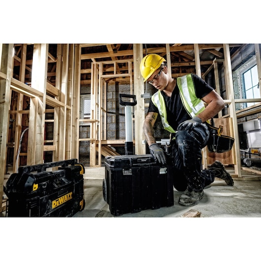 A construction worker wearing a yellow hard hat and reflective vest is kneeling next to DEWALT tool storage boxes in a building under construction. The tool storage boxes are black with yellow accents, and wooden framing is visible in the background.