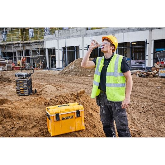 DEWALT yellow toolbox on construction site, worker in vest and helmet drinks water next to it.