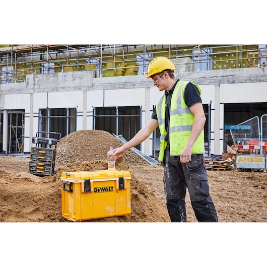 Yellow DEWALT toolbox on construction site with a worker in safety gear placing a bottle on it.