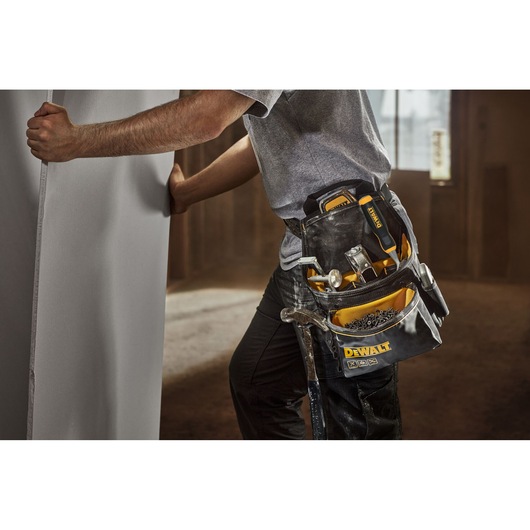 A person wearing a DEWALT tool belt holds a large panel. The tool belt contains various hand tools, including a hammer, screwdrivers, and fasteners. The scene is set indoors, suggesting a construction or installation environment.