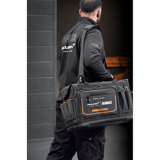 Person carrying a black DEWALT McLaren tool bag with orange accents in an industrial setting.