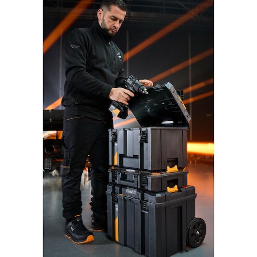 Person with blurred face holding a power tool near stacked DEWALT toolboxes with wheels in an indoor setting.