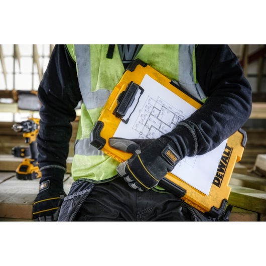 A person wearing work gloves and a high-visibility vest is holding a yellow DEWALT clipboard case with architectural drawings attached, at a construction site.