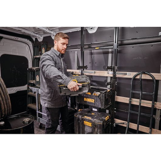 A person with a blurred face is organizing DEWALT tool storage boxes inside a utility van. The boxes are stacked and labeled