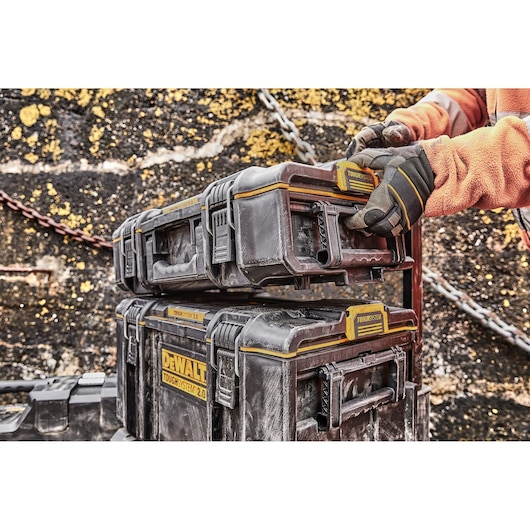 Gloved hands stacking DEWALT ToughSystem 2.0 toolboxes against a rough outdoor wall.