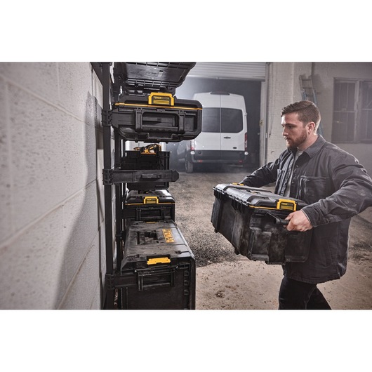 A person is holding a DEWALT ToughSystem storage box and placing it onto a rack with several other similar storage boxes in