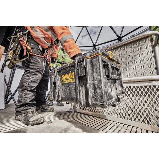 A person wearing work gear and safety harness lifts a DEWALT ToughSystem 2.0 tool box on an industrial site with metal