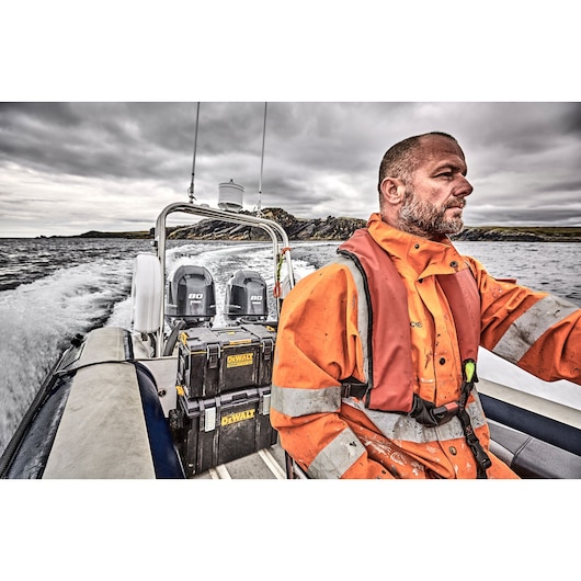 A person wearing an orange safety jacket and life vest is operating a motorboat on the water. Two DEWALT DWST83294-1